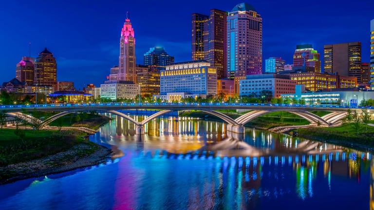 Columbus skyline illuminated at night with colorful buildings and city lights reflecting on the Scioto River below a curved bridge